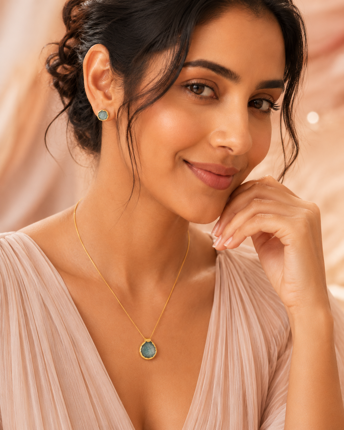 A woman wearing a delicate gold necklace with a round amazonite pendant and matching small stud earrings, styled with a pleated light pink top against a soft, warm beige background.